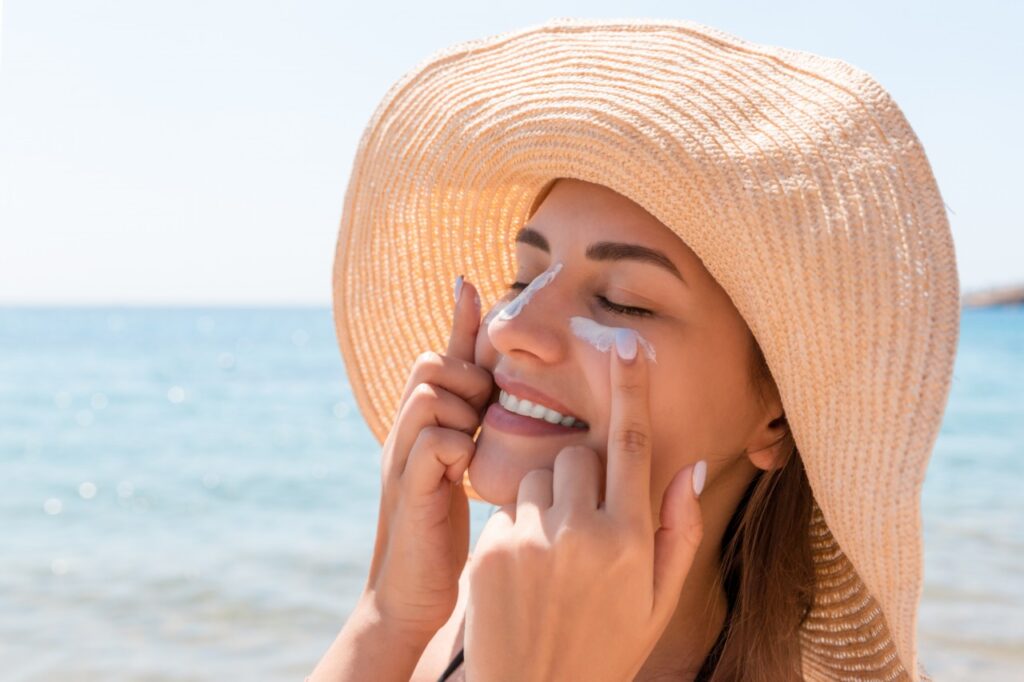 chica con sombrero en la playa aplicándose crema en el contorno de los ojos